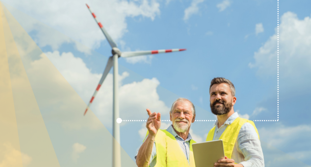 A pair of workers hold a clipboard in front of a wind turbine.